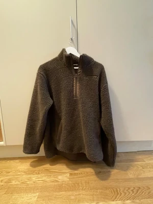 Brun teddytröja med halv dragkedja - Mysig brun teddytröja med halv dragkedja framtill och hög krage. Tröjan har lång ärm och är tillverkad i ett fluffigt, stickat material som ger en avslappnad och trendig look. Perfekt för kyliga dagar när du vill vara både varm och snygg.