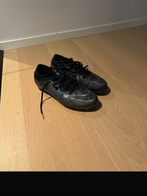 Svarta Nike fotbollsskor med snörning - Säljer ett par svarta Nike fotbollsskor med klassisk snörning och låg profil. Skorna har en sportig design med vita detaljer och är tillverkade i syntetmaterial för bra grepp och känsla på planen. Perfekta för dig som vill ha stilrena och funktionella skor till fotbollen.