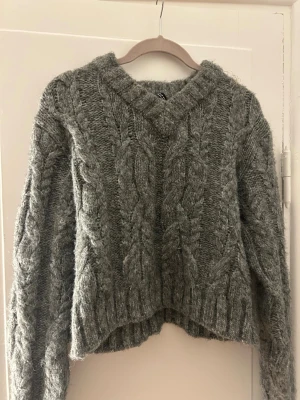 Grå kabelstickad tröja med v-ringning - Mysig grå stickad tröja med kabelstickat mönster och v-ringad hals. Tröjan har långa ärmar och ribbade muddar vid ärmslut och nederkant. Perfekt för kyligare dagar och styling! Superbra skick! Säljer då den inte kommit till användning! ☺️