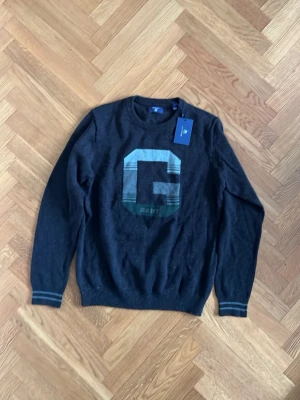 Mörkgrå stickad tröja från GANT - Snygg mörkblå stickad tröja från GANT. Helt ny och oanvänd, därför med prislappen kvar. Stort snyggt G i mitten.