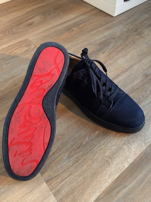 Marinblåa sneakers från Christian Louboutin - Snygga marinblåa sneakers från Christian Louboutin med klassisk röd sula och svarta skosnören. Skorna har en låg profil och är tillverkade i mocka för en stilren och exklusiv look. Perfekta för dig som vill sticka ut med en ikonisk detalj. Skorna är lite skadade som syns på bild 4 och 5 men det är inget som märks när man använder dem. Köpta här på plick.