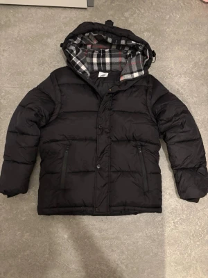 Burberry jacka - Svart puffer dunjacka med huva och rutigt foder i grått, vitt och rött. Jackan har dragkedja och knappar framtill samt två fickor med dragkedja. Perfekt för kalla dagar och har en klassisk, oversized look.