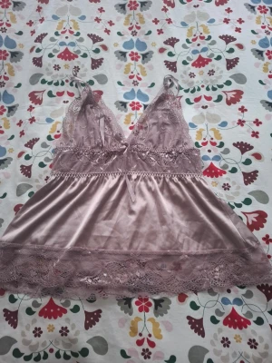 Rosa spetsnattlinne med satin - Sött nattlinne i ljusrosa satin med spetsdetaljer och tunna axelband. Djup V-ringning och dekorativ rosett framtill. Spets längs byst och nederkant ger en romantisk känsla. Perfekt för mysiga kvällar hemma.