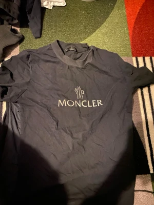 Svart Moncler t-shirt med logga - Snygg svart t-shirt från Moncler med klassisk logga tryckt i vitt på bröstet. T-shirten har rund hals och korta ärmar, perfekt för en clean och stilren look. Tillverkad i mjuk bomull som känns skön mot huden.