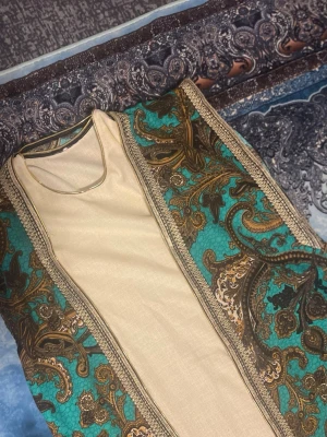 Turkos och beige mönstrad väst - Unik väst i turkos och beige med detaljerat paisleymönster i brunt och guld. Västen har dekorativa guldfärgade kanter och öppen front. Perfekt för dig som vill sticka ut med en färgstark och elegant look.