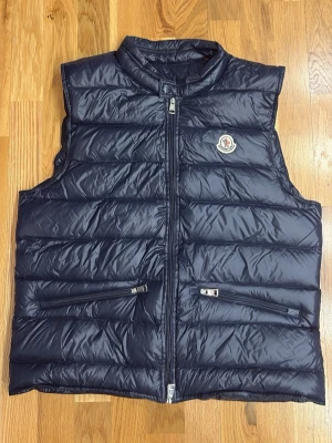 Mörkblå väst moncler - Snygg mörkblå dunväst från Moncler med klassisk quiltad design och hög krage. Västen har två dragkedjeförsedda fickor framtill och ikonisk Moncler-logga på bröstet. Tillverkad i glansigt material som ger en exklusiv känsla. Size 3/L