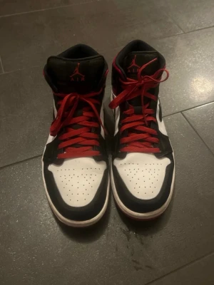 Nike Air Jordan 1 High svart/vit/röd - Säljer ett par Nike Air Jordan 1 High sneakers i svart, vitt och rött. Klassisk design med svart swoosh, röda skosnören och detaljer, samt perforerad tå. Ovandel i läder och hög siluett som ger en ikonisk streetwear-look.