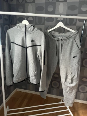 Gråa Nike Tech Fleece mjukisbyxor L - Snygg grå hoodie från Nike med hel dragkedja och svart logga på bröstet. Hoodien har huva, svarta detaljer längs sömmarna och är tillverkad i mjukt sweatshirtmaterial. Perfekt för en sportig och avslappnad stil. Tröjan är storlek M och byxorna storlek L, men passar M