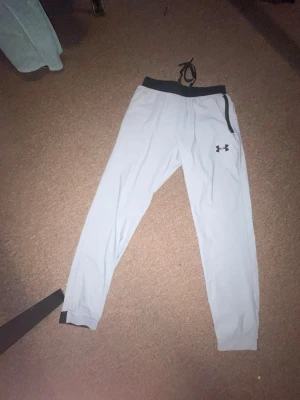 Grå mjukisbyxor från Under Armour - Snygga grå mjukisbyxor från Under Armour med svart resår i midjan och dragsko. Byxorna har smal passform, muddar vid bensluten och en svart logga på ena benet. Perfekta för chill eller träning.