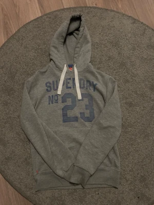 Grå Superdry hoodie med tryck - Snygg grå hoodie från Superdry med blått tryck 'No 23' på bröstet och vita dragsnören i huvan. Klassisk känguruficka framtill och ribbade muddar. Perfekt för en avslappnad streetstyle-look.