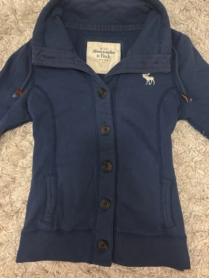 Blå kofta från Abercrombie & Fitch - Mörkblå kofta med knappar framtill från Abercrombie & Fitch. Har hög krage, två fickor och en vit älglogga broderad på bröstet. Mjuk bomullskvalitet och ribbade muddar vid ärmslut och nederkant. Står storlek L men passar perfekt på S ♥️