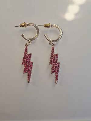 Guldörhängen med rosa blixtar - Snygga örhängen i guldton med hängande blixtar täckta av små rosa stenar. Blixtarna har en cool, edgy form och örhängena har en rund, räfflad bygel. Perfekta för dig som vill sticka ut med färg och unik design.