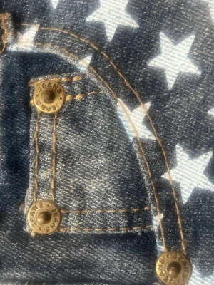 Jeansshorts med stjärnor och ränder - Snygga jeansshorts med fransig kant, ena sidan har vita stjärnor på blå denim och andra sidan har blå och vita ränder. Klassisk femficksmodell med nitar och knapp i mässing. Perfekta för en cool och avslappnad stil. Står på lappen att det är M men väldigt små i storleken