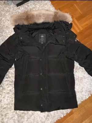 Canada goose svart - Säljer en svart dunjacka från Canada Goose med pälskantad huva och tydlig logotyp på ärmen. Jackan har en klassisk, vadderad design med knappar och dragkedja framtill. Perfekt för kalla vinterdagar och riktigt snygg till alla outfits.