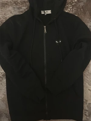 Svart hoodie från Comme des Garçons Play - Svart hoodie med dragkedja från Comme des Garçons Play. Klassisk modell med huva, snörning och det ikoniska hjärta med ögon på bröstet. Tillverkad i mjuk bomull och har långa ärmar samt fickor framtill.