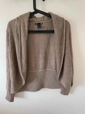 Beige ribbad kofta från H&M - Mysig beige kofta från H&M med ribbad struktur och öppen front. Koftan har långa ärmar och en avslappnad passform, perfekt att slänga över en topp. Tillverkad i mjukt stickat material som känns skönt mot huden.