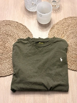 Olivgrön långärmad tröja Polo Ralph Lauren - Snygg olivgrön långärmad tröja från Polo Ralph Lauren i mjuk bomull. Klassisk passform med rund halsringning och den ikoniska vita logobroderingen på bröstet. Perfekt basplagg för en clean och stilren look.