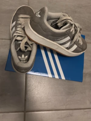 Grå Adidas Campus sneakers - Snygga grå Adidas Campus sneakers med klassiska vita ränder på sidorna och vit sula. Ovandelen är i mocka och skorna har lågt skaft. Perfekta för dig som gillar retrostil och streetwear. Kommer med gråa skosnören.