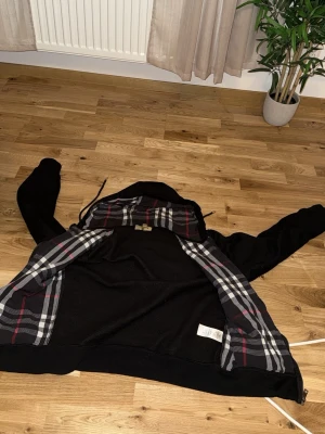 Svart Burberry Hoodie  -  Säljer en svart  Burberry hoodie med dragkedja och huva i storlek M, men passar också S. Insidan av huvan har ett klassiskt rutigt mönster i svart, vitt och rött. Hoodien har fickor framtill och justerbara snören i huvan. Perfekt för en chill och stilren look.