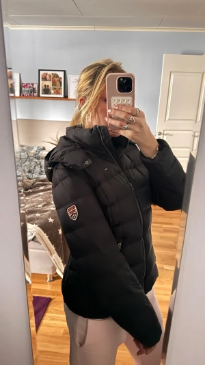 Svart pufferjacka Tommy Hilfiger - Snygg svart pufferjacka från Tommy Hilfiger med huva och dragkedja framtill. Jackan har två fickor med dragkedja och ett broderat märke på ärmen. Perfekt för kalla vinterdagar och riktigt skön att bära.