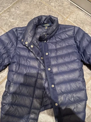 Mörkblå dunjacka från Polo Ralph Lauren - Snygg mörkblå dunjacka från Polo Ralph Lauren med hög krage och tryckknappar. Jackan har quiltad design och diskret logga på bröstet. Perfekt för kalla vinterdagar och riktigt stilren look.