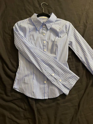 Blårandig skjorta från Bershka  - Snygg blå skjorta med vita vertikala ränder. Skjortan har klassisk krage, långa ärmar och knäppning framtill. Figurnära passform och tillverkad i ett mjukt bomullsmaterial som känns bekvämt hela dagen. Den är figursydd och har utsvävningar vid underarmen.