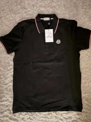 Svart Moncler pikétröja med röd/vit kant - Snygg svart pikétröja från Moncler med klassisk krage och knäppning framtill. Tröjan har röda och vita detaljer på krage och ärmslut samt Moncler-logga på bröstet. Perfekt för en clean och stilren look. Helt oanvänd, L sitter som M