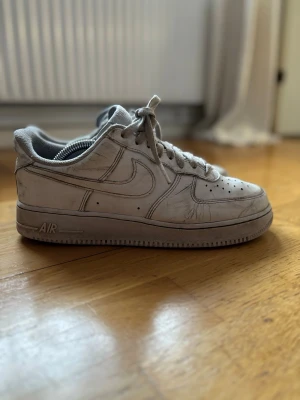 Nike Air Force 1 vita sneakers - Klassiska vita Nike Air Force 1 sneakers med lågt skaft. Skorna har den ikoniska swoosh-loggan på sidan och AIR-detalj på sulan. Tillverkade i läder med textilfoder och snörning framtill. Perfekta för en streetwear-look. Lite skadad i hälen med något hål där