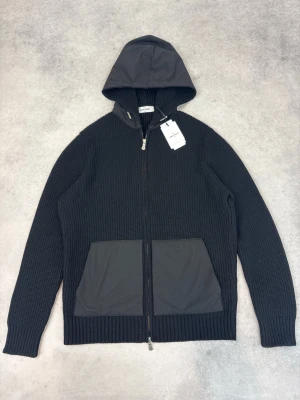 Svart hoodie från Gran Sasso - Stilren svart hoodie från Gran Sasso med stickad struktur och dragkedja framtill. Huva och fickor i kontrasterande material ger en modern touch. Perfekt för dig som gillar en clean look med lite edge. Två stora fickor framtill och ribbade muddar.