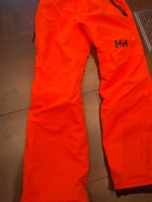 Överdragsbyxor helly hansen - Helly Hansen teknbyxor i orange färg. I nyskick / mycket fint skick. Vattentäta och vindtäta, perfekta för skidåkning, friluftsliv eller vardag vintertid. Inga fläckar eller skador.
