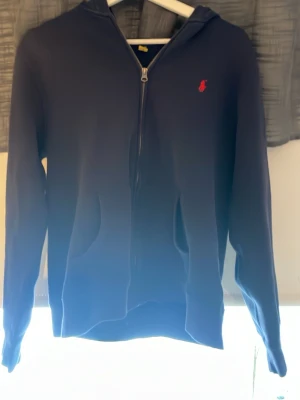 Mörkblå hoodie från Ralph Lauren - Mörkblå hoodie från Ralph Lauren med dragkedja framtill och klassisk röd logga broderad på bröstet. Tröjan har huva och två fickor framtill. Tillverkad i mjukt material som passar perfekt för en avslappnad stil.