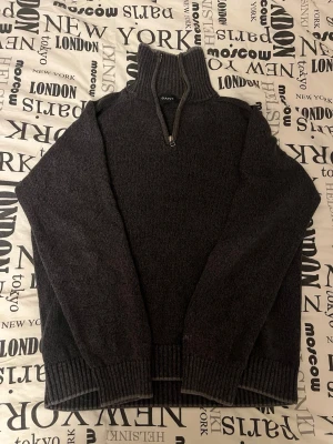 Mörkgrå stickad halvzip från GANT - Mörkgrå stickad tröja från GANT med halv dragkedja och hög ribbad krage. Tröjan har långa ärmar och ribbade muddar vid ärmslut och nederkant. Perfekt för lager på lager och en stilren look.