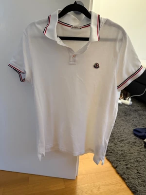 Vit Moncler piké med röd detalj - Vit pikétröja från Moncler med klassisk krage och röda samt marinblå ränder på krage och ärmslut. Liten Moncler-logga på bröstet. Tillverkad i bomull och har korta ärmar. Perfekt för en clean och sportig look.