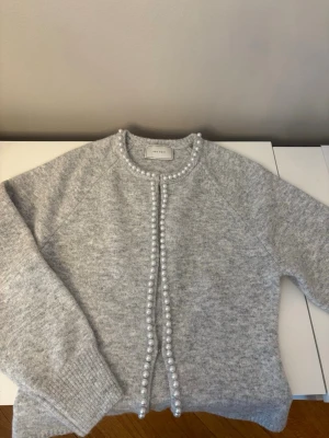 Grå kofta från neo noir -  grå kofta från neo noir med pärldetaljer längs knäppningen och runt halsen. Mjuk och mysig, perfekt till lager-på-lager. Rund halsringning och långa ärmar. Materialet är en stickad blandning som känns skön mot huden.