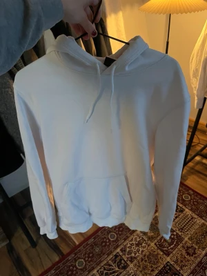 Vit hoodie med huva och magficka - Säljer en stilren vit hoodie med klassisk huva och dragsnören. Hoodien har en stor magficka framtill och ribbade muddar vid ärmslut och nederkant. Perfekt för en chill och avslappnad look. Materialet känns mjukt och bekvämt, troligtvis bomullsblandning.