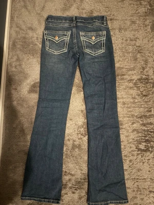 Blå bootcut jeans med kontrastsömmar - Snygga blå bootcut jeans med markerade kontrastsömmar och fickor med lock och knapp bak. Klassisk femficksmodell med låg midja och lätt utsvängda ben. Perfekta för en avslappnad och trendig look. Använt en gång💕