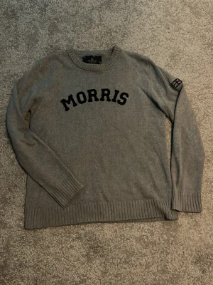 Grå stickad tröja från Morris - Snygg grå stickad tröja från Morris med svart text på bröstet och en patch med brittiska flaggan på ärmen. Tröjan har rund halsringning, ribbade muddar och är tillverkad i mjukt stickat material. Perfekt för dig som gillar klassisk och stilren design.
