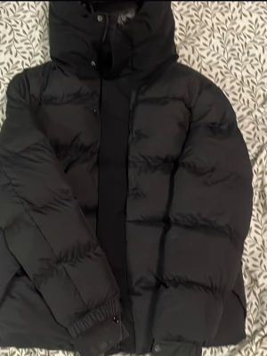Svart moncler  pufferjacka med luva ny - Säljer en svart pufferjacka med stor luva och fuskpälsdetalj. Jackan har en quiltad design, dragkedja och tryckknappar framtill samt Moncler-logga på ärmen. Perfekt för kalla vinterdagar och riktigt snygg till streetwear-stilen.
