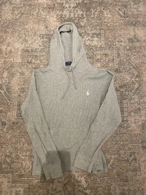 Grå hoodie från Polo Ralph Lauren - Snygg grå hoodie från Polo Ralph Lauren i ribbad bomull med klassisk huva och dragsnören. Tröjan har en broderad vit logga på bröstet och en stor magficka. Perfekt för en chill och avslappnad stil.