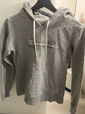 Grå hoodie från Champion - Snygg grå hoodie från Champion med tryckt logga på bröstet och klassisk känguruficka. Tröjan har huva med vita dragsnören och ribbade muddar vid ärmslut och nederkant. Tight passform. Använd men felfri