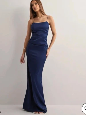 Marinblå långklänning med tunna band - Elegant marinblå långklänning med smala axelband och figurnära passform. Klänningen har öppen rygg och är tillverkad i ett glansigt, mjukt material som ger en lyxig känsla. Perfekt för dig som ska gå på bal.