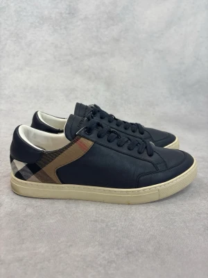 Burberry mörkblåa sneakers med rutmönster - Vi har dessa skor i storlek 41 och 42. Snygga svarta sneakers från Burberry i läder med klassiskt beige, svart och rött rutmönster på sidan. Skorna har vita sulor och svarta skosnören. Perfekt för dig som vill ha en stilren och exklusiv look med en touch av brittisk design.