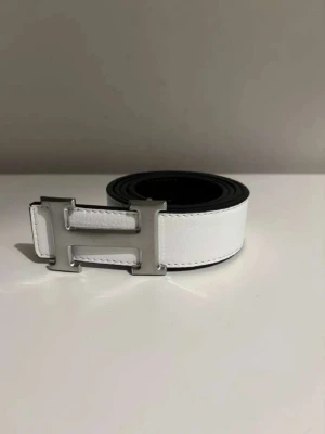 Vitt skärp med H-spänne i skinn - Snyggt vitt skärp från  med klassiskt H-format spänne i silverfärg. Bältet är gjort i skinn och har en stilren, minimalistisk look som passar till många outfits. Perfekt accessoar för dig som gillar exklusiva detaljer.