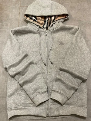 Grå zip hoodie från Burberry - Snygg grå hoodie med dragkedja från Burberry. Klassisk huva med justerbara snören och ikoniskt rutigt foder i beige, svart och rött. Liten diskret Burberry-logga broderad på bröstet. Mjuk bomullsblandning och ribbade muddar vid ärmar och nederkant.
