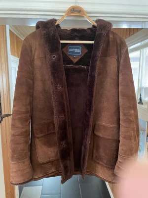 Brun fårskinnskappa East West - Säljer en brun fårskinnskappa från East West med mjukt foder och stora fickor framtill. Kappan har huva och är tillverkad i mocka med fluffigt innerfoder. Perfekt för kalla dagar och ger en retrovibe till din outfit.