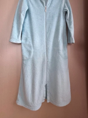 Ljusblå fleece-morgonrock med dragkedja - Mysig, lång  ljusblå morgonrock i mjuk fleece med diskret mönster, långa ärmar och praktiska fickor. Dragkedja framtill gör den enkel att ta på och av. Perfekt för att hålla värmen under kyliga morgnar.