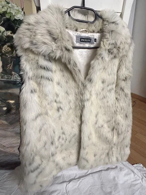 Pälsväst från Isolde - Snygg ljus pälsväst från Isolde med mjuk fuskpäls och diskreta mörkare inslag. Västen har en klassisk krage och silkeslen insida. Perfekt för att lyfta din outfit med en trendig touch. Storlek XL men sitter bra på mig som använder S/M så den är väldigt liten i storleken 