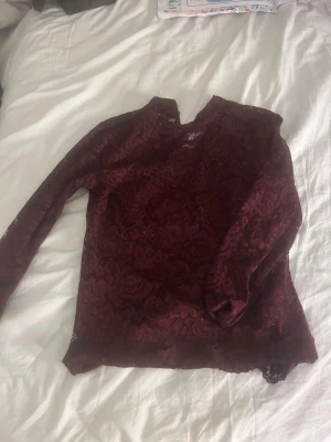 Vinröd spetsblus med lång ärm - Vinröd blus i spets med lång ärm och hög hals. Blusen har ett vackert blommigt spetsmönster och är lätt transparent. Perfekt för dig som vill ha en elegant och trendig topp med det lilla extra.