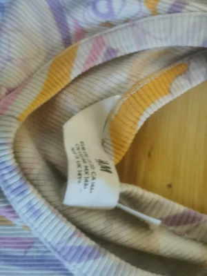 Mönstrad t-shirt från H&M - Cool t-shirt från H&M med färgglatt psykedeliskt mönster i lila, rosa, gult och vitt. T-shirten har korta ärmar och är gjord i ribbad bomull, vilket ger en skön och trendig känsla. Perfekt för dig som gillar unika prints och vill sticka ut.