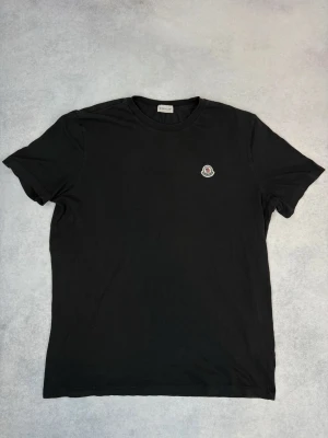 Svart Moncler t-shirt med logga - Säljer en svart t-shirt från Moncler med rund hals och liten broderad Moncler-logga på bröstet. Klassisk och stilren modell i mjuk bomull, perfekt till jeans eller shorts. Enkel men snygg detalj som lyfter hela outfiten.
